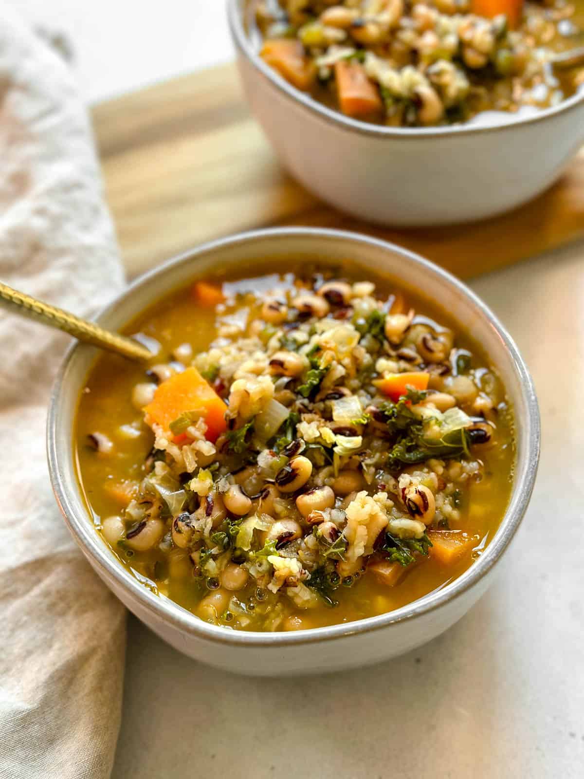 A gray bowl filled with vegan black-eyed pea soup with a golden spoon and tan napkin on a white background. A second bowl on a wooden platter is in the background.