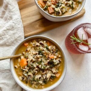 Bowl of black-eyed pea soup in a light gray bowl served with a spoon. Off to the side is a festive pink drink with ice and a sprig of rosemary in it.