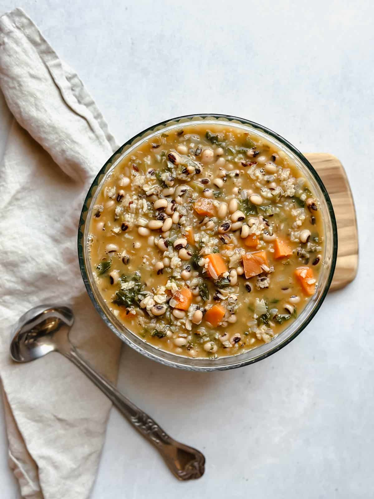 Large bowl of vegan Black-Eyed Pea Soup with Kale and Rice in a glass bowl with a ladle and tan napkin off to the side.