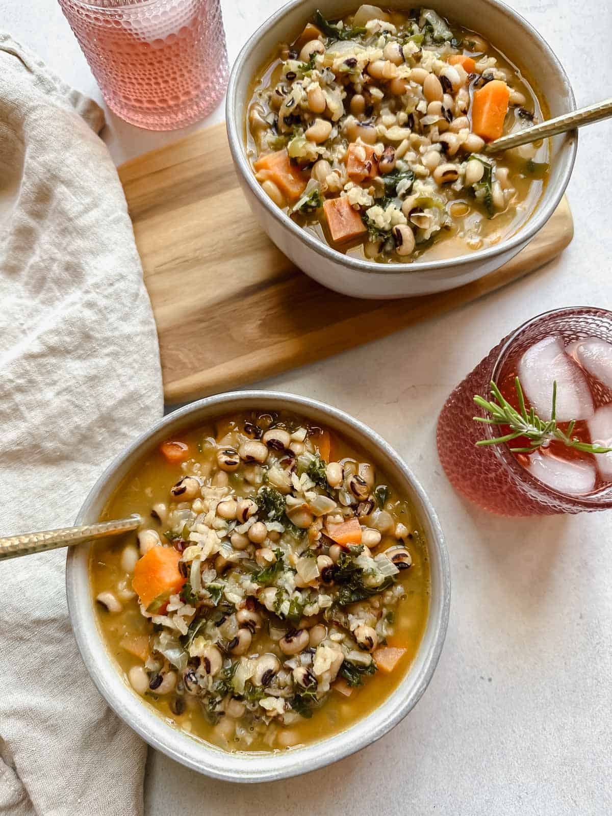 Two bowl of black-eyed pea soup served in light gray bowls each with a spoon. Along side there is a tan napkin and two festive pink drinks with ice and a sprig of rosemary in them. One of the bowls is sitting on a wooden serving platter. All on white background.