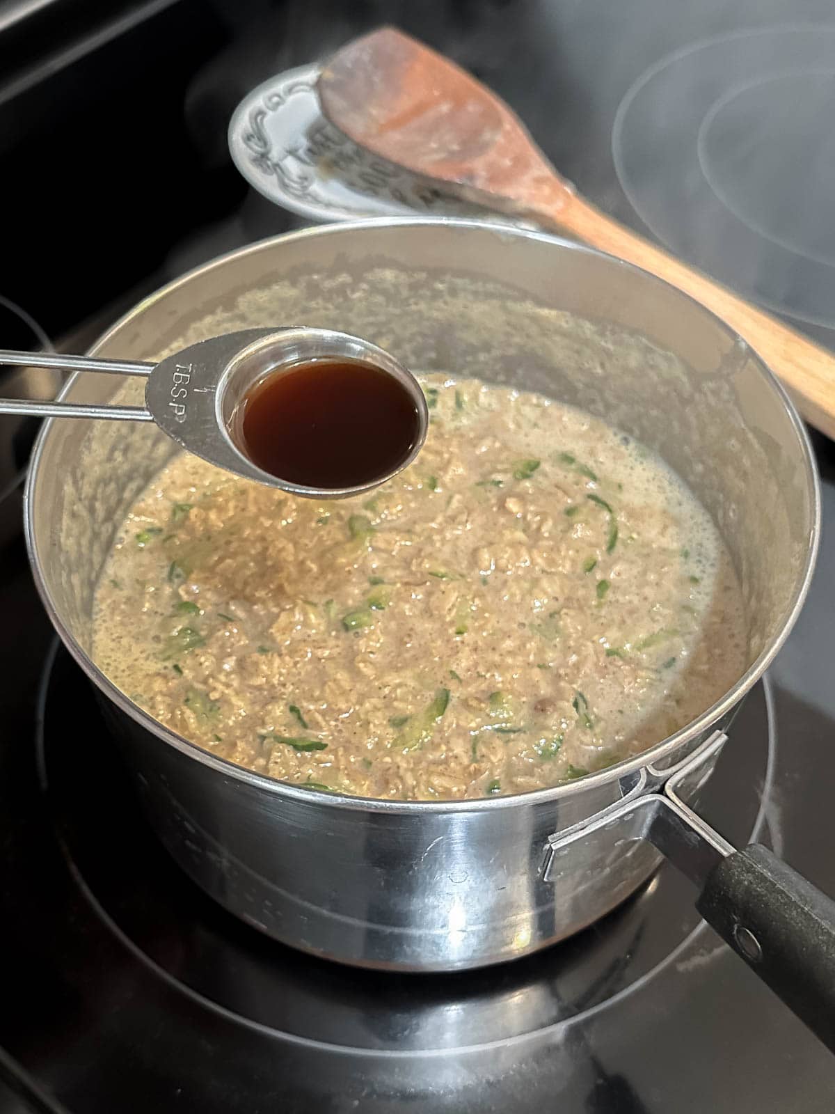 A stainless steel tablespoon filled with vanilla extract over a pot of oatmeal on the stove