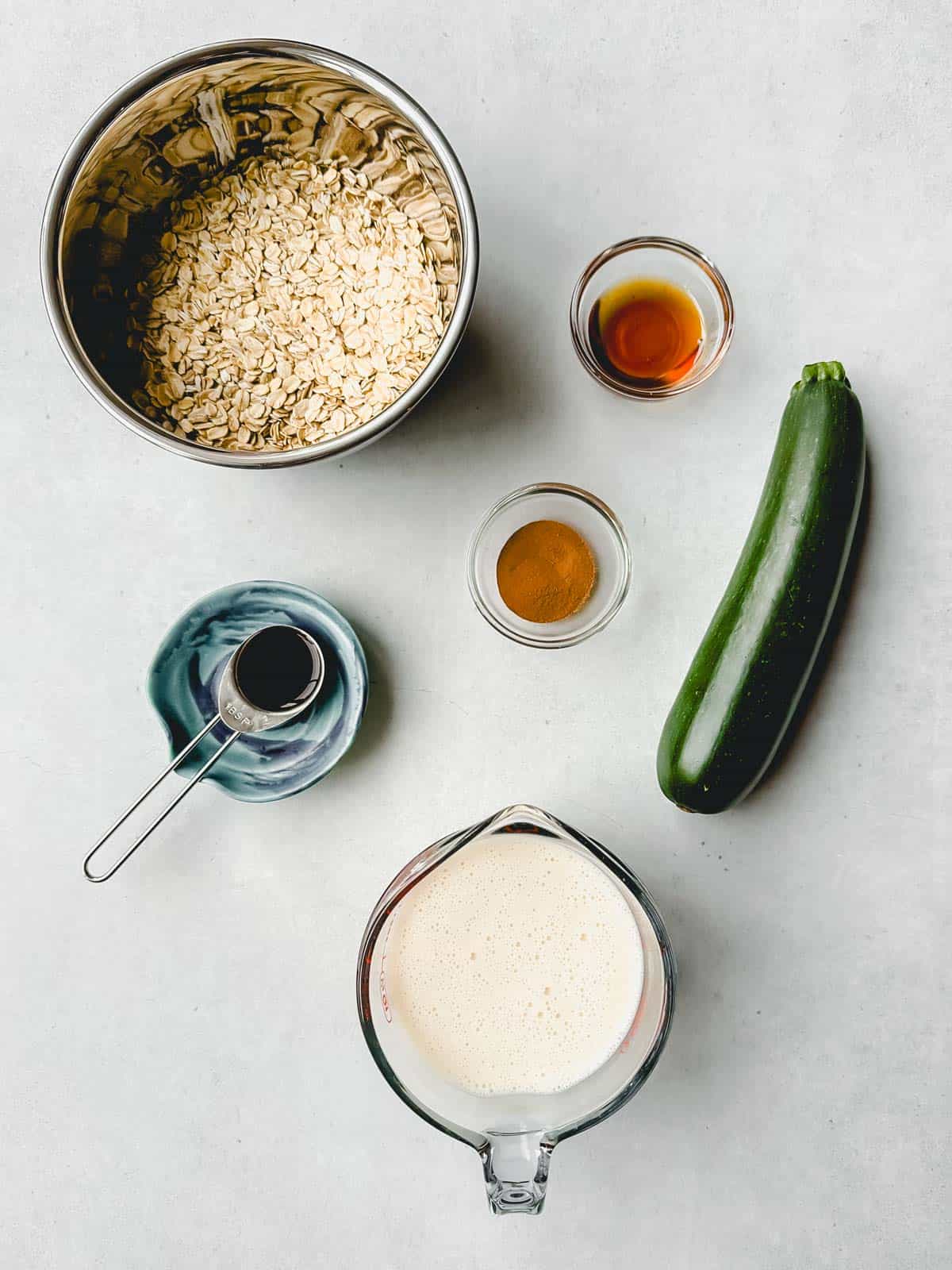 Individual ingredients to make zucchini oatmeal: rolled oats in stainless steel bowl, a whole zucchini, soy milk in measuring cup, and maple syrup, cinnamon and vanilla extract in small containers.