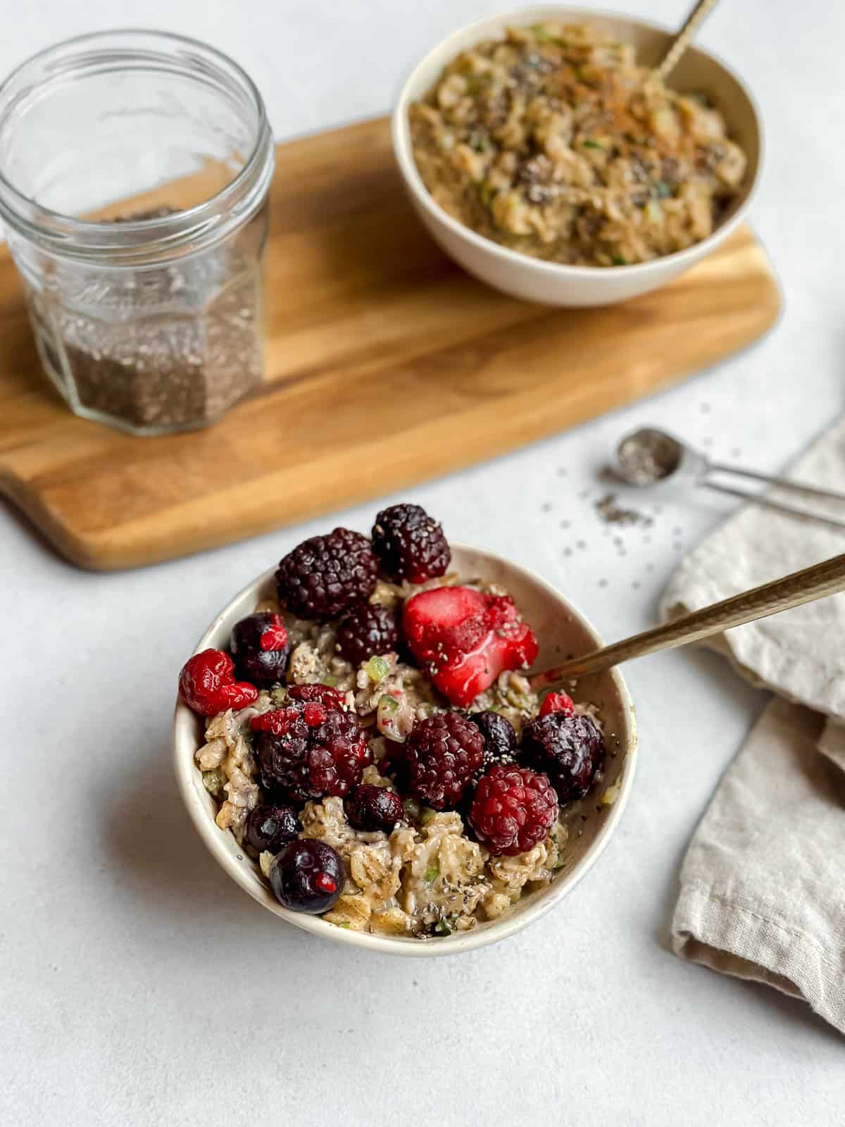 Vegan zucchini oatmeal topped with berries and chia seeds served in a white bowl with a spoon. An additional bowl in the background and a glassware of chia seeds sitting on a wooden serving platter.