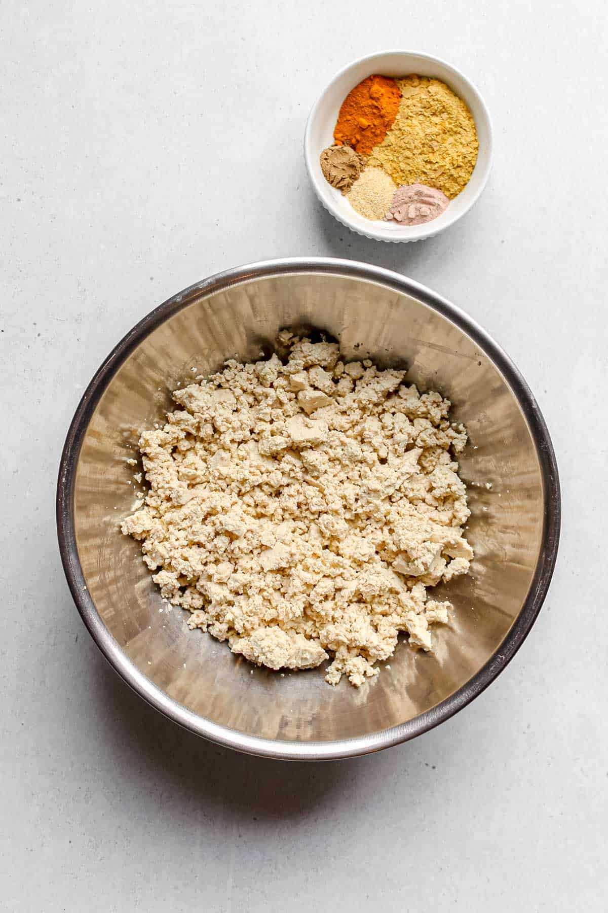 Crumbled firm tofu in large stainless steel bowl and spices in small white bowl
