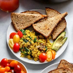 Tofu scramble with onion, spinach, and mushroom served on white plate with toast, grape tomatoes, and sliced avocado.