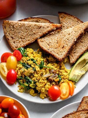 Tofu scramble with onion, spinach, and mushroom served on white plate with toast, grape tomatoes, and sliced avocado.