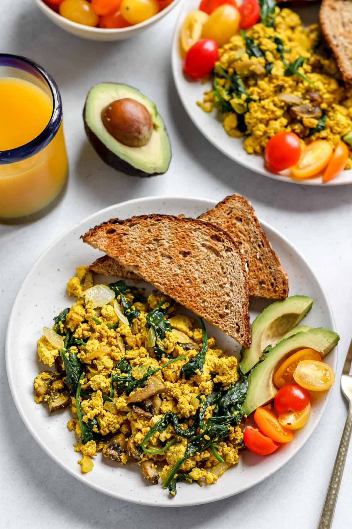 Tofu scramble with spinach and mushroom served on white plate with toast, grape tomatoes, sliced avocado and a glass of orange juice in a glass with a blue rim.