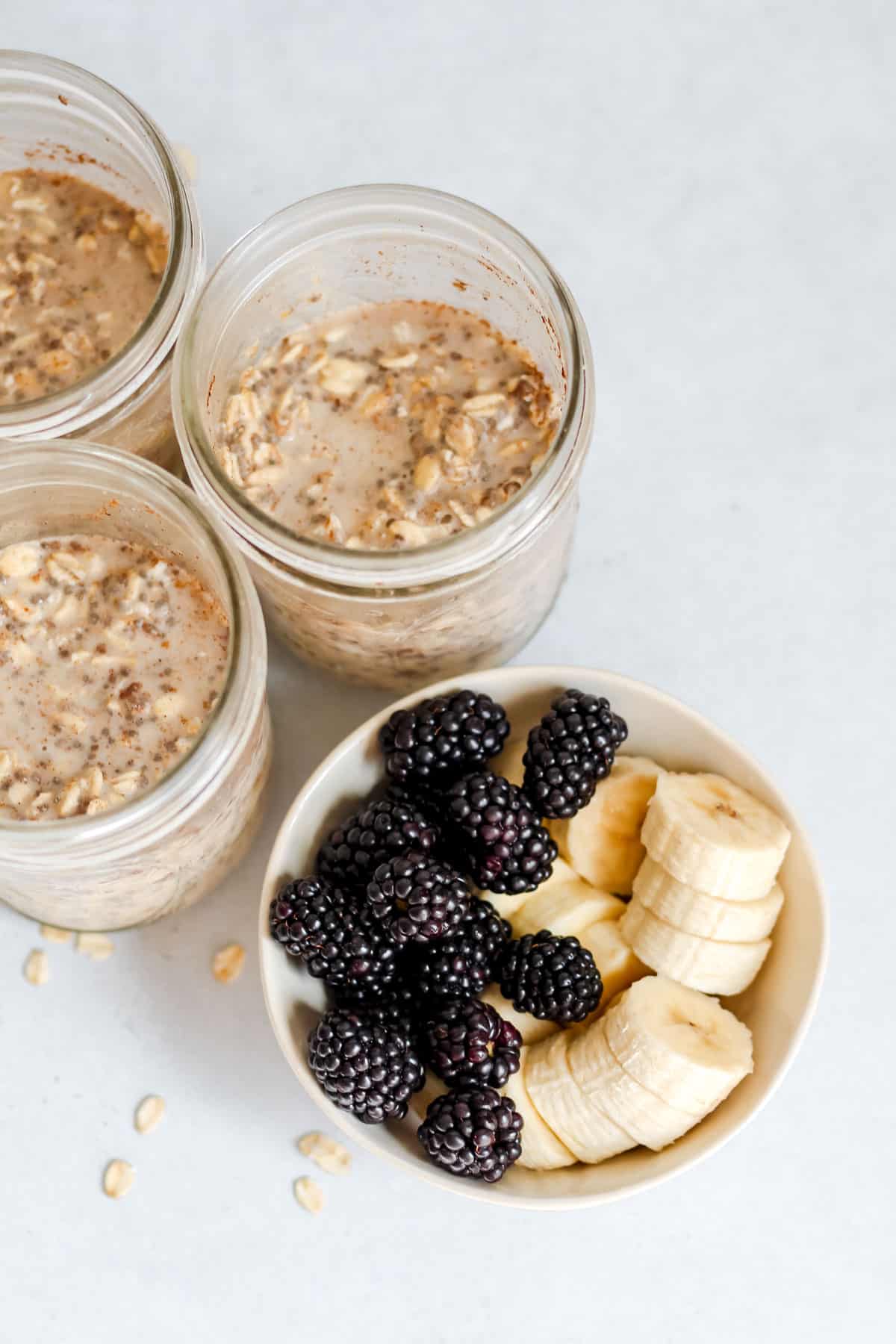 Blackberries and sliced banana in a white bowl along with 3 mason jars full of overnight oats