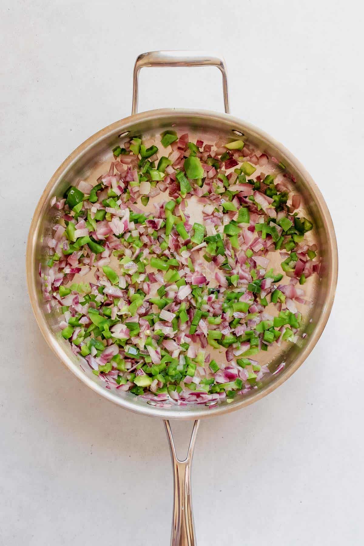 Sautéed green bell pepper, jalapeño and red onion in a stainless steel skillet on a white background.