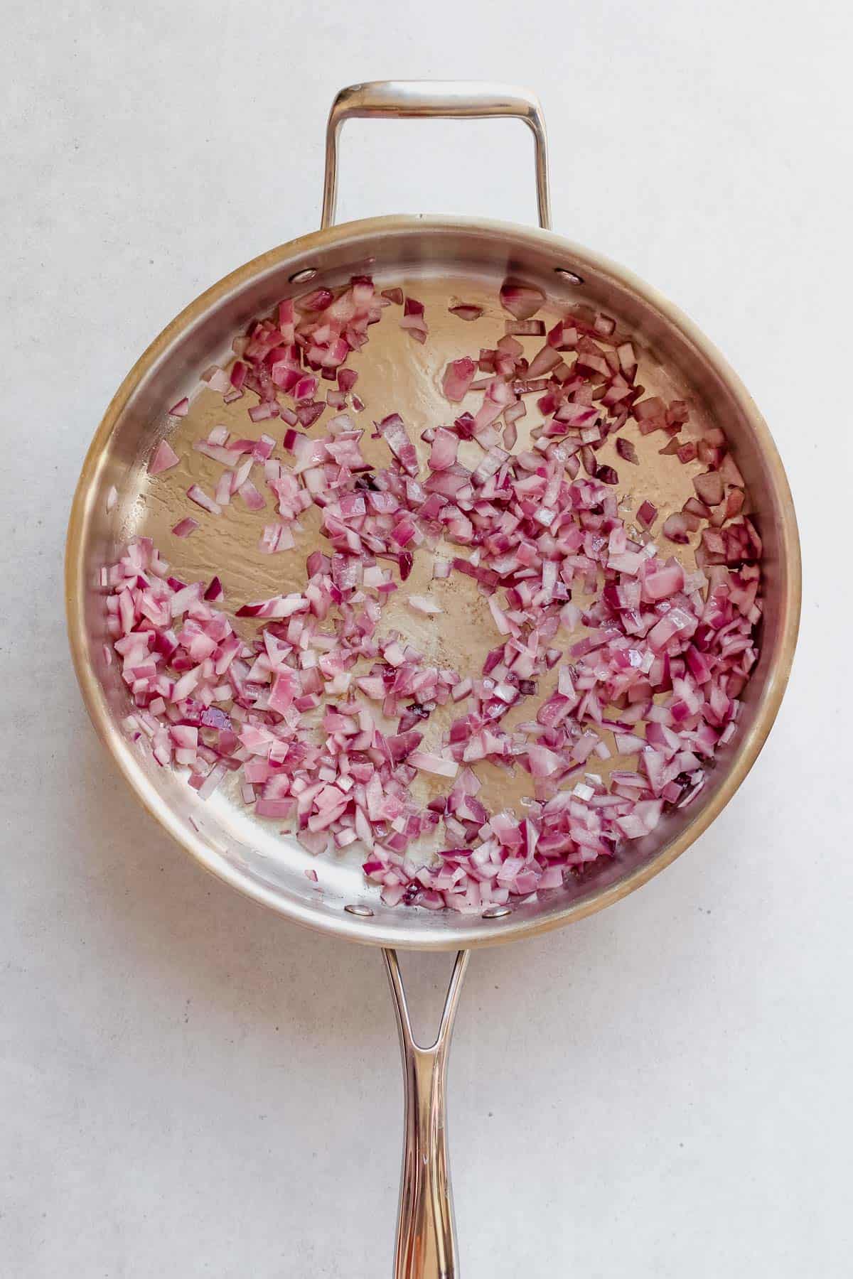 Sautéed red onion in a stainless steel skillet on a white background.