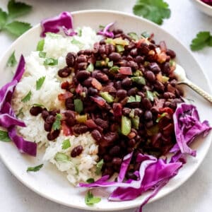 Cooked spicy black beans served with white rice and garnished with cilantro and purple cabbage on white plate.