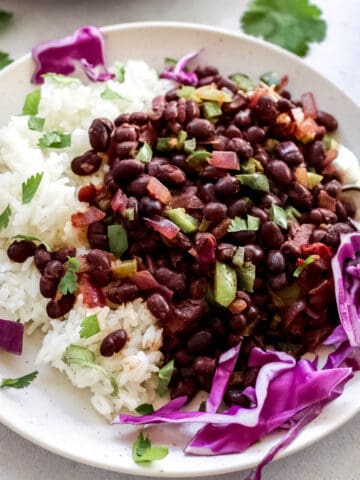 Cooked spicy black beans served with white rice and garnished with cilantro and purple cabbage on white plate.