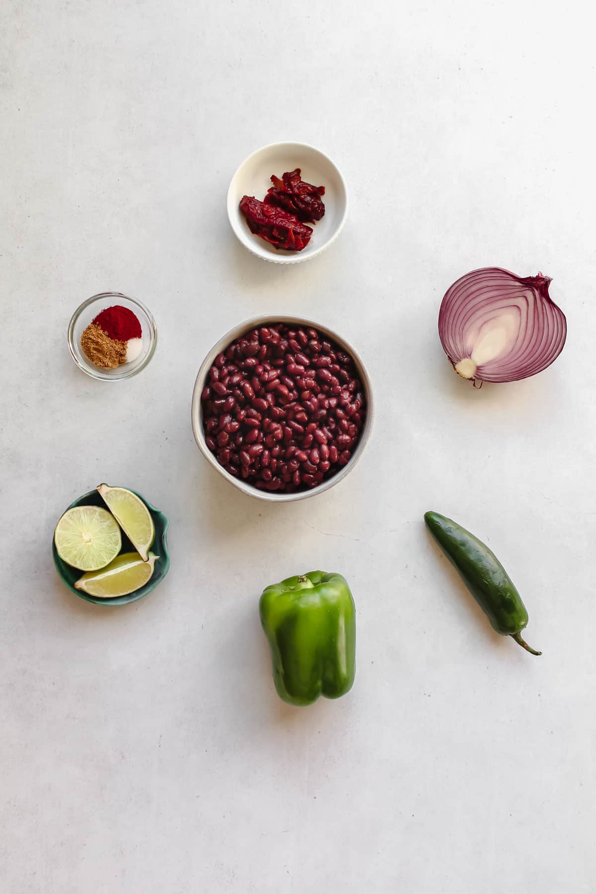Individual Ingredients: black beans in a bowl, a half of a red onion, 1 green bell pepper, 1 jalapeño pepper, spices, chipotle peppers, and lime wedges
