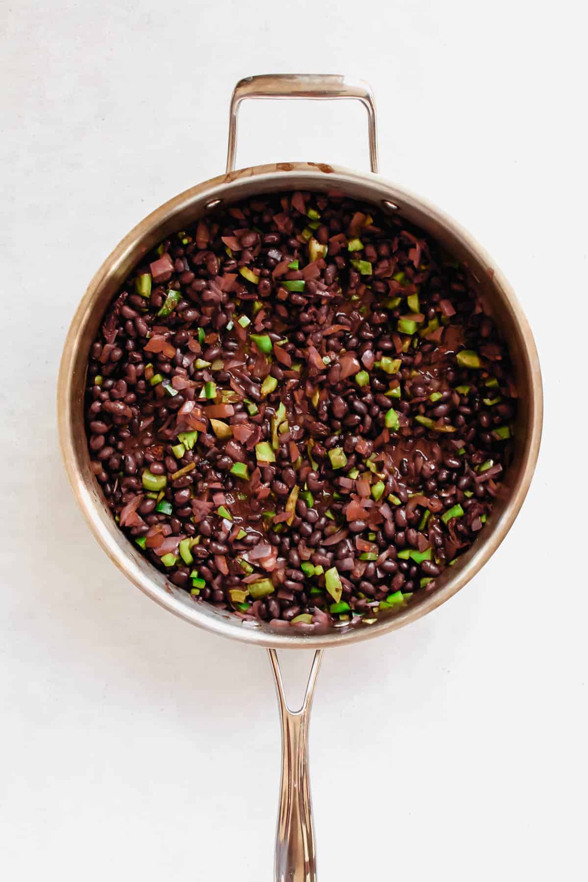 Spicy black beans mixed with sautéed green bell pepper, jalapeño and red onion in a stainless steel skillet on a white background.
