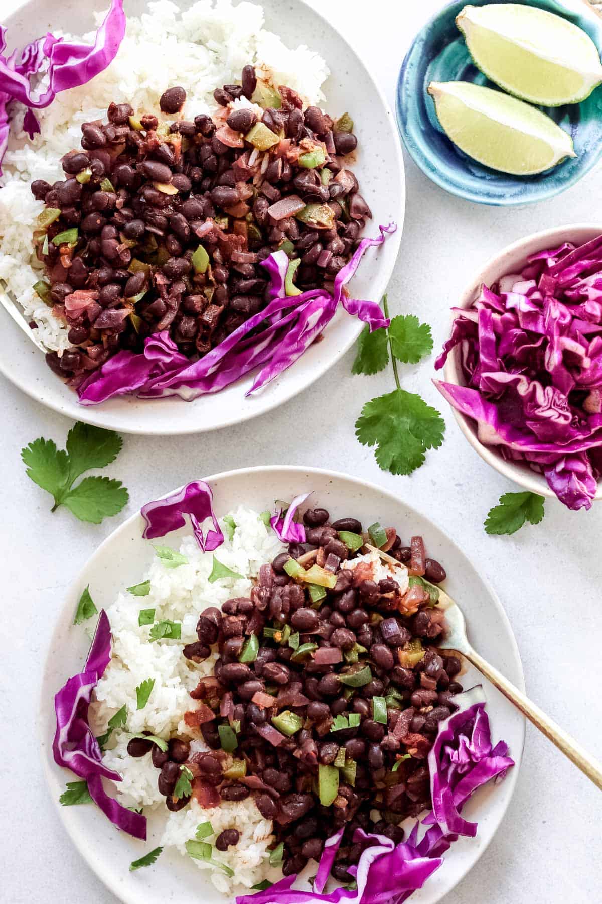 Cooked spicy black beans plated with rice and garnished with cilantro and purple cabbage. Extra lime wedges in a small blue bowl and extra purple cabbage in a small white bowl.