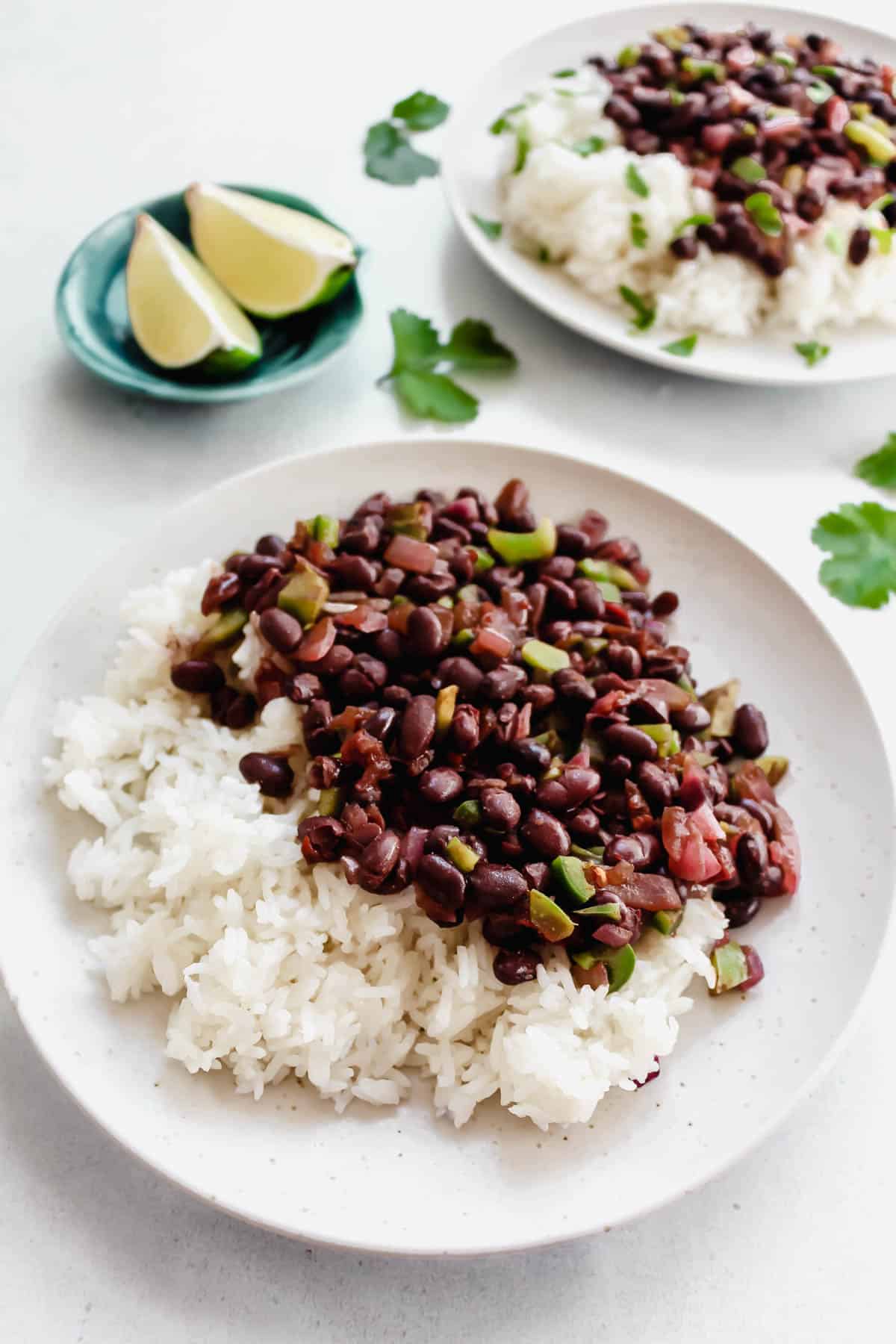 Spicy black beans served with white rice and cilantro on white plates. 2 lime wedge in a small blue bowl.