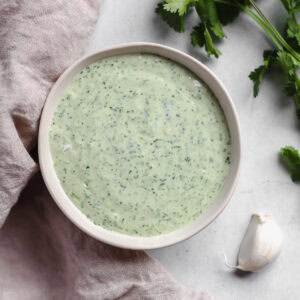 Creamy cilantro lime crema in a white bowl along side a tan cloth, a garlic clove, and sprigs of fresh cilantro