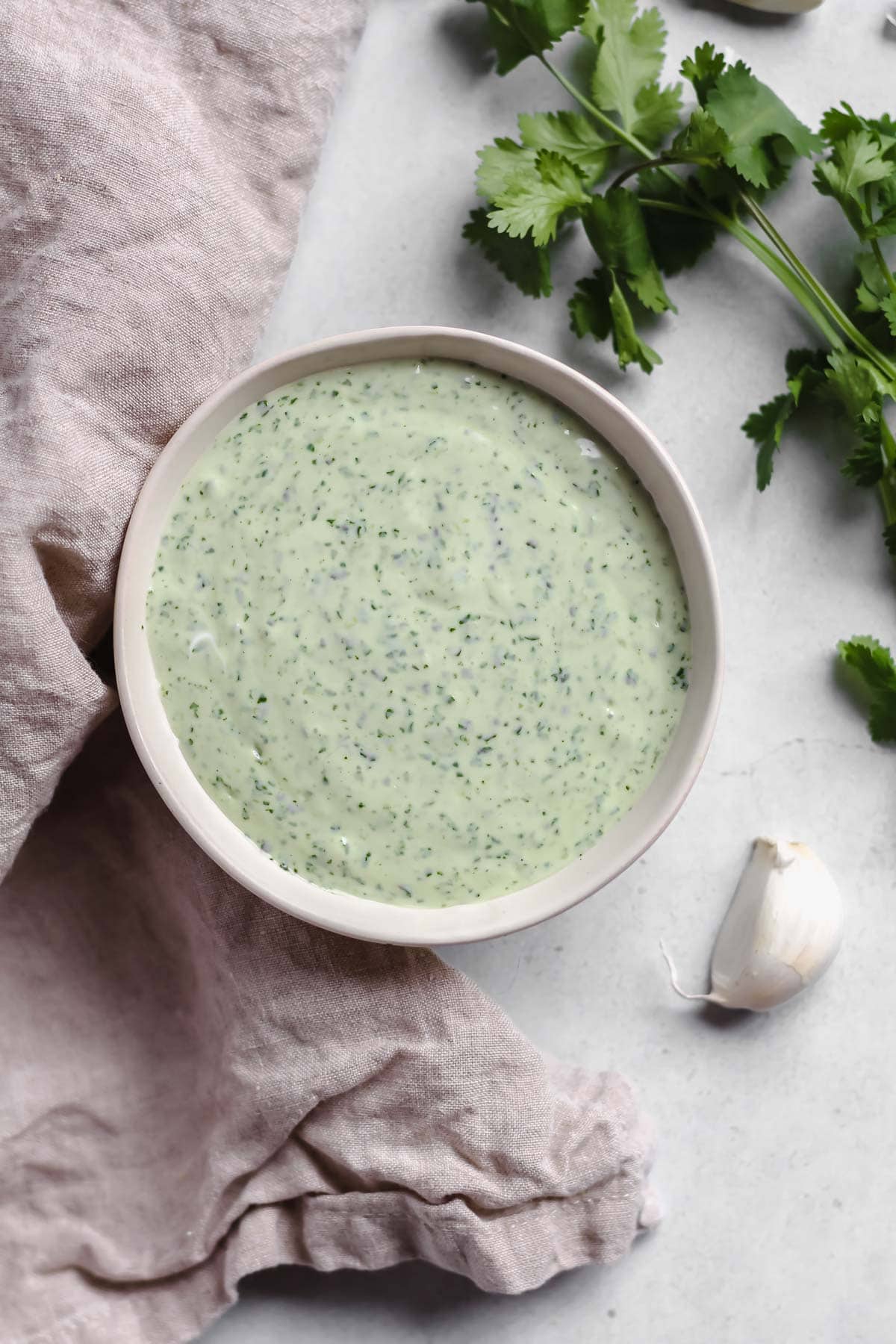 Creamy cilantro lime crema in a white bowl along side a tan cloth, a garlic clove, and sprigs of fresh cilantro