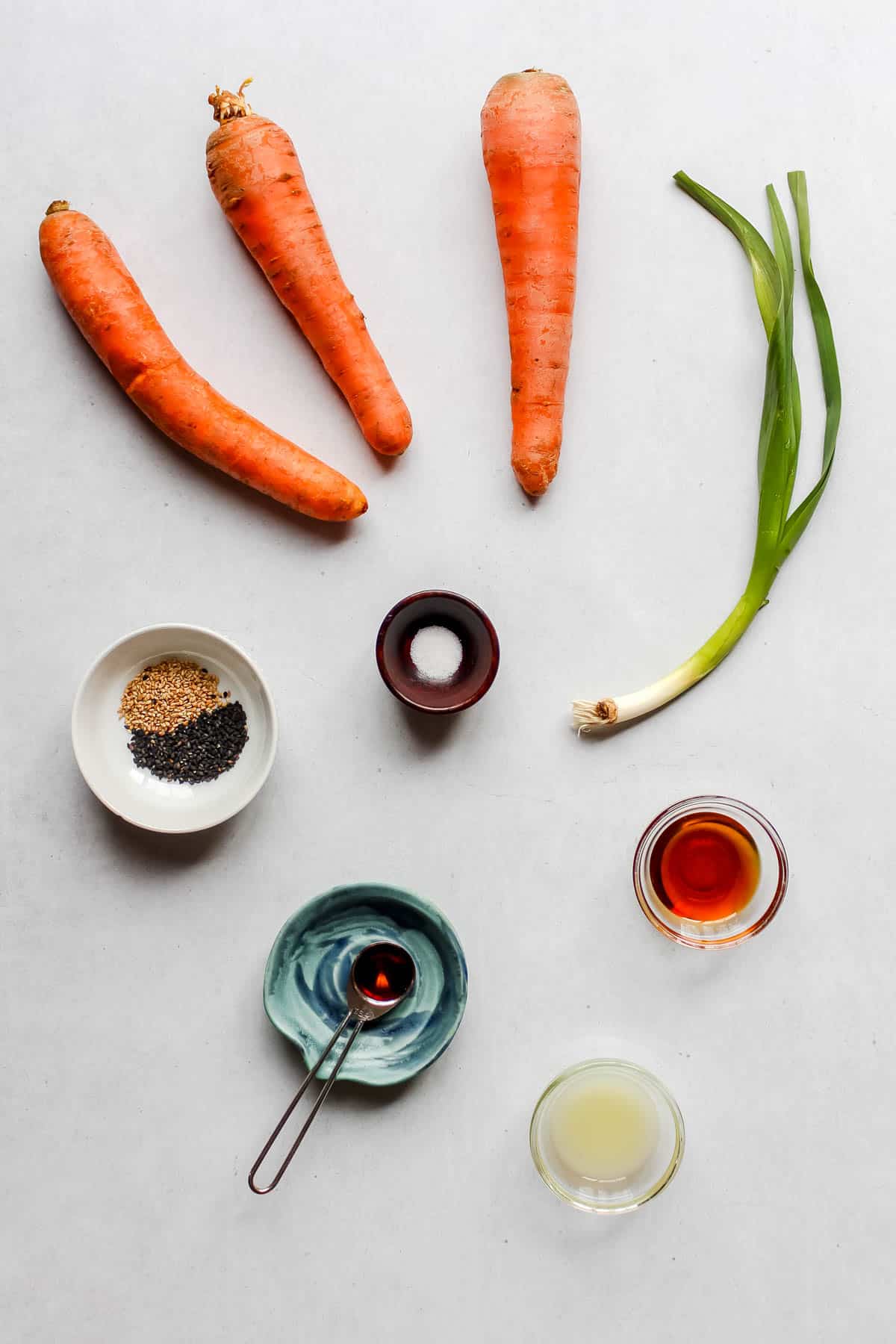 Individual ingredients to make a raw carrot salad on a white back ground: 3 large carrots, 1 green onion, sesame seeds in small white bowl, salt in small brown bowl, sesame oil, lime juice, and maple syrup