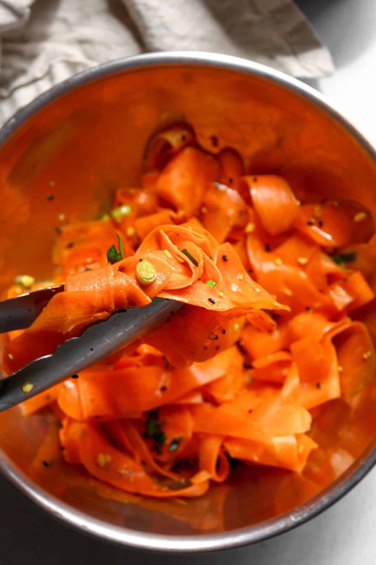 Mixing the dressing, sesame seeds, and green onions into the carrot salad in a stainless steel bowl with tongs.