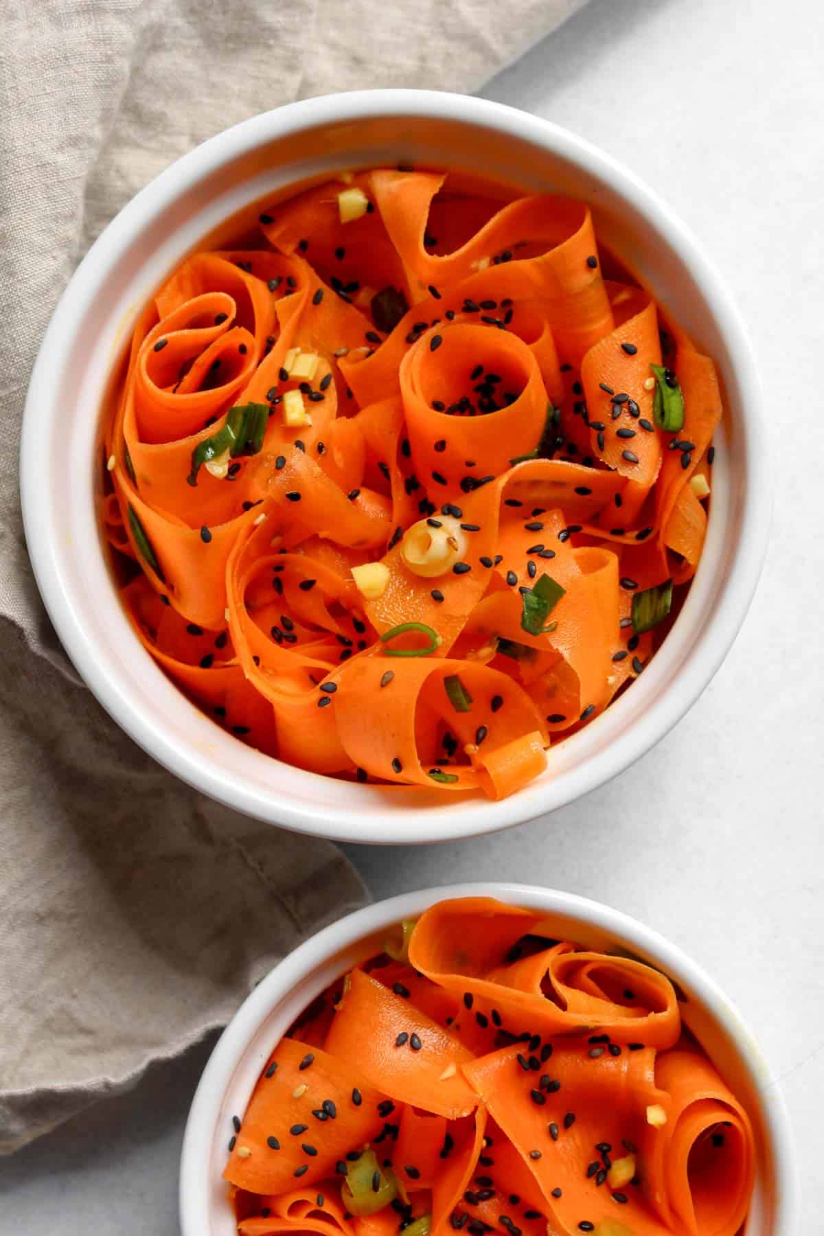 Carrot ribbons served in two white bowls topped with black sesame seeds and sliced green onion