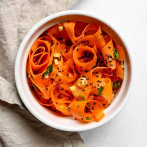 Raw carrot salad served in a white bowl along side a tan napkin on a white background.
