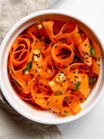 Raw carrot salad served in a white bowl along side a tan napkin on a white background.