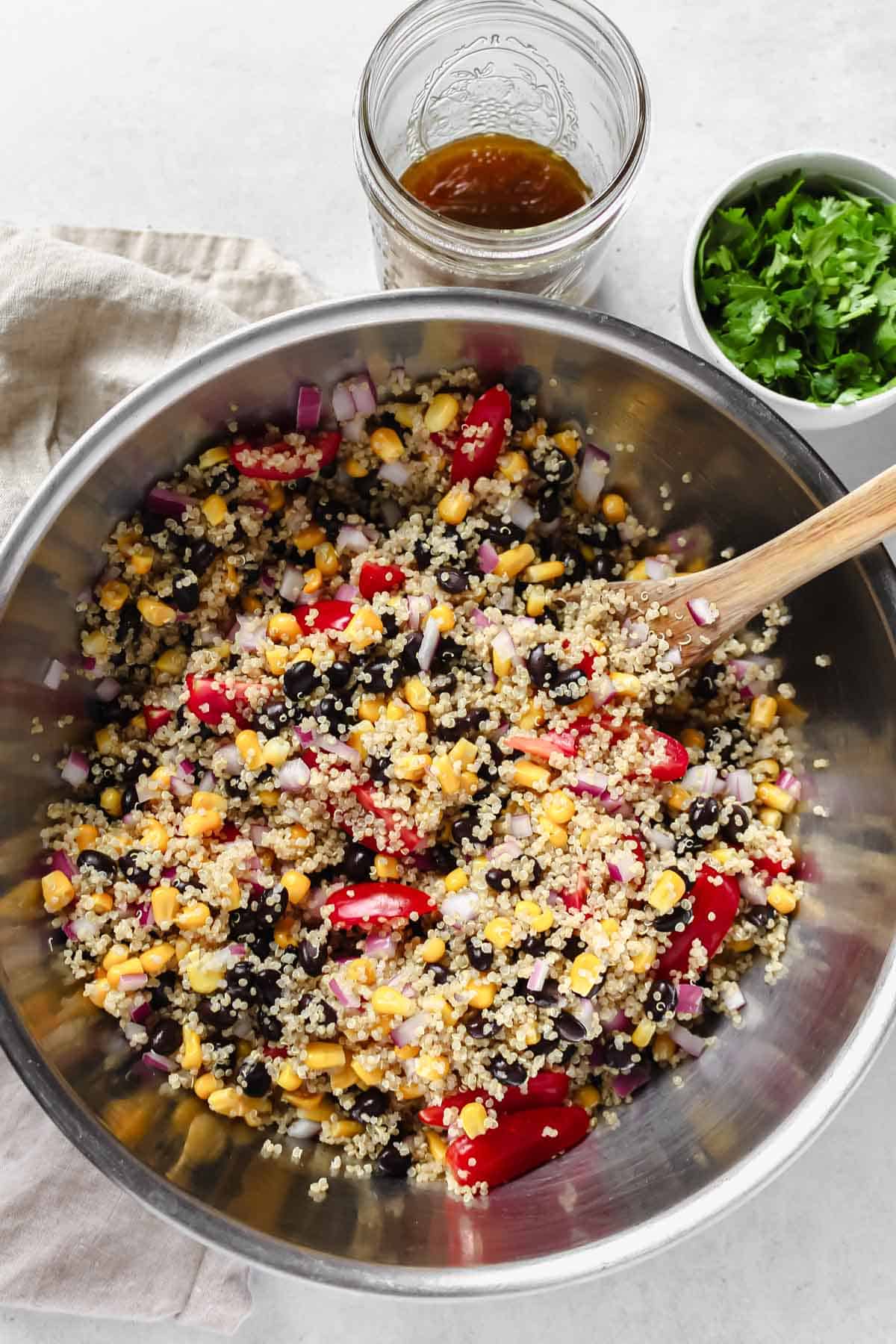 Combining cooked quinoa, black beans, corn, tomatoes, and red onion in a mixing bowl with a wooden spoon, alongside a glass mason jar of dressing and a cup of cilantro.