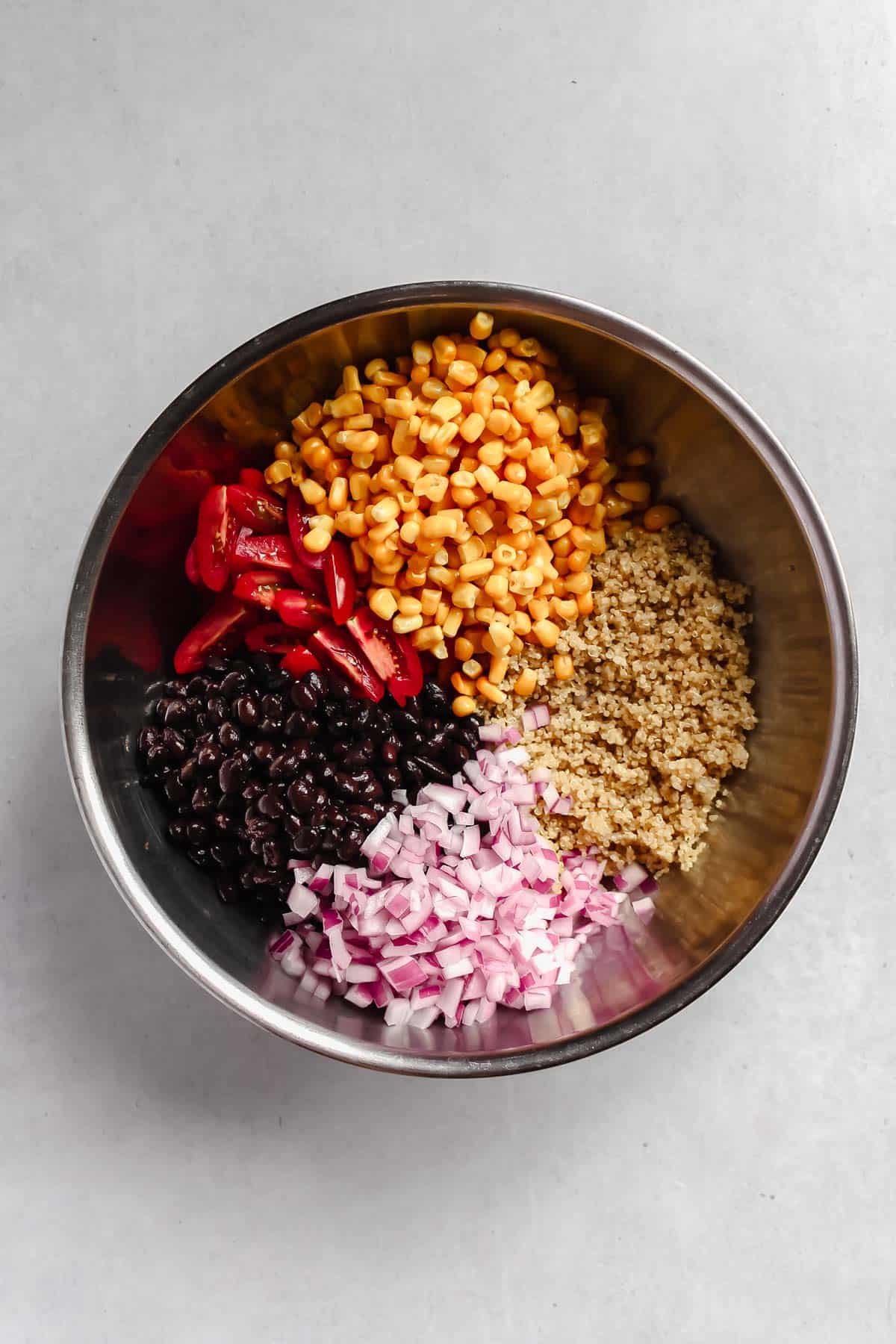 Cooked quinoa, black beans, corn, tomatoes, and red onion in a stainless steel mixing bowl