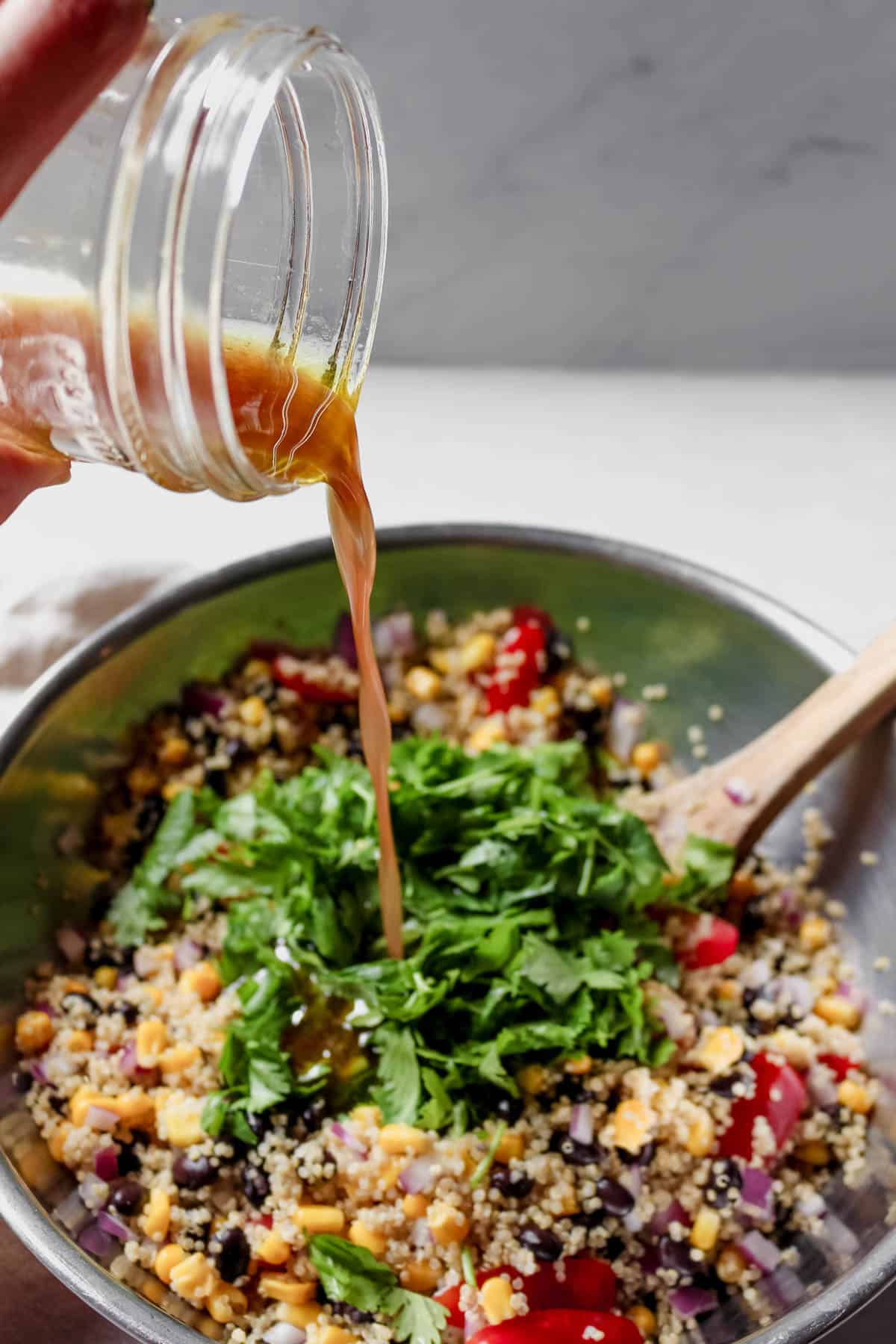 Pouring the dressing out of a glass mason jar into the mixing bowl full of quinoa salad ingredients