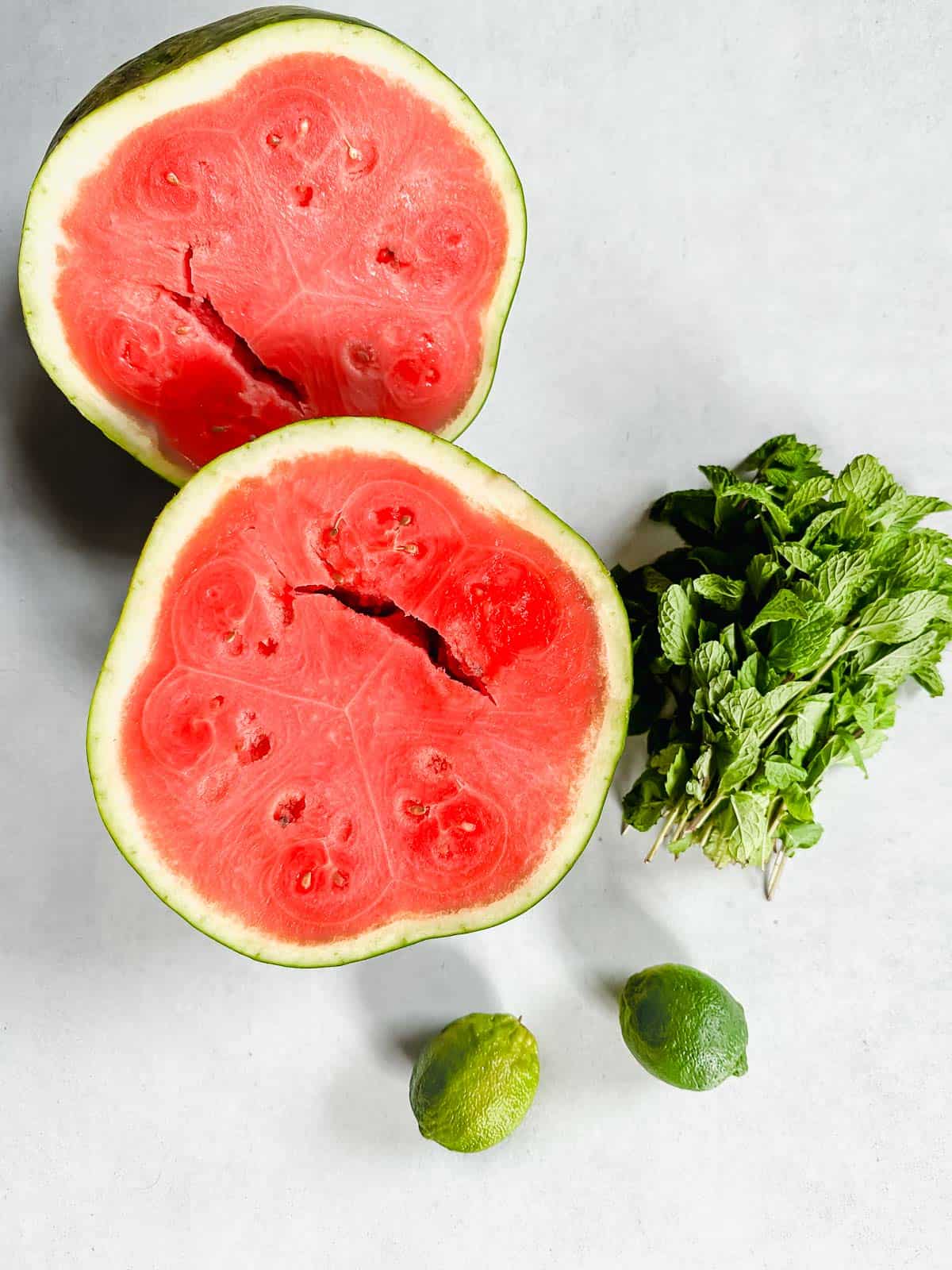 A cut in half watermelon, two whole limes, and a bunch of mint