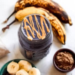 Blueberry Banana Peanut Butter Smoothie with peanut butter drizzled on top, surrounded by ingredients like bananas, blueberries, and flaxseed.