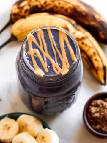 Blueberry Banana Peanut Butter Smoothie with peanut butter drizzled on top, surrounded by ingredients like bananas, blueberries, and flaxseed.