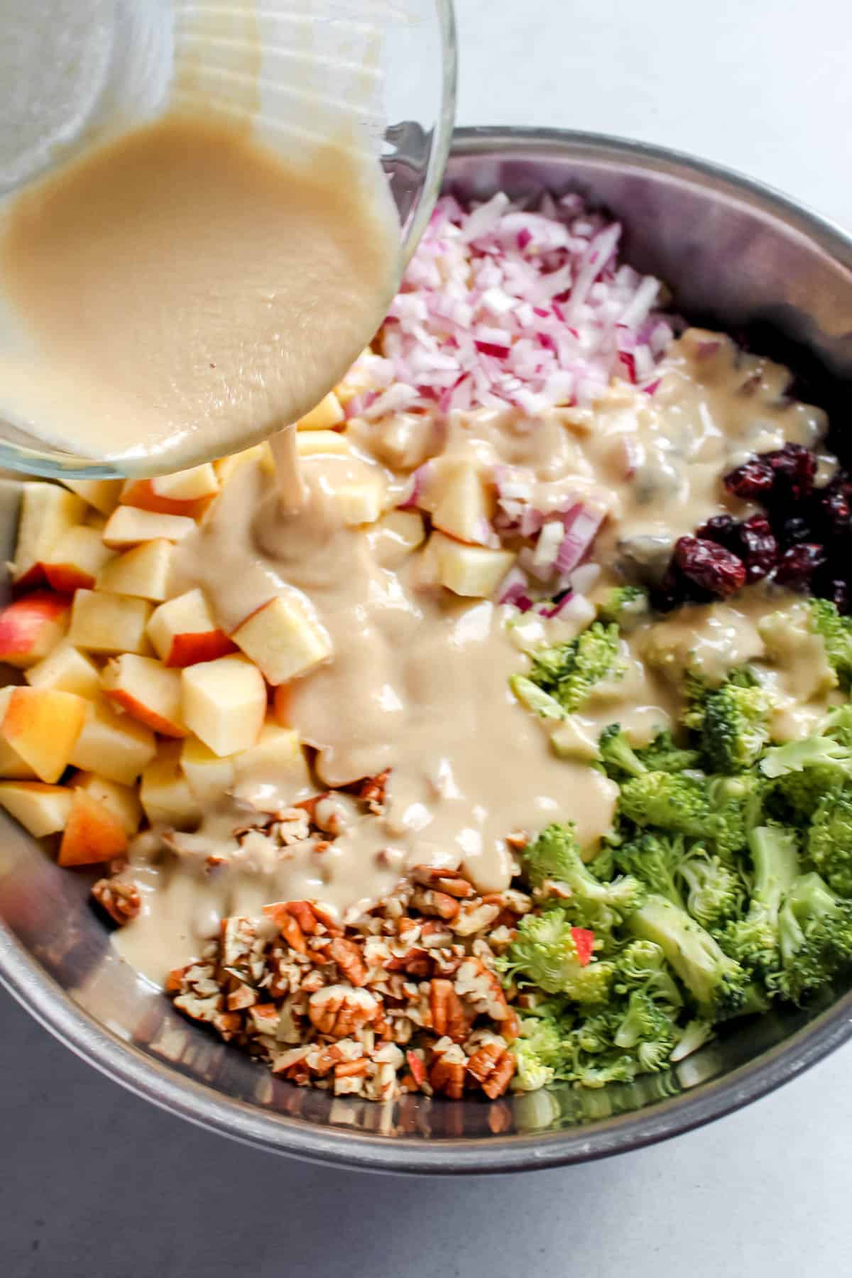 Tahini dressing being poured on the chopped salad ingredients that are in a large stainless steel bowl on a white background