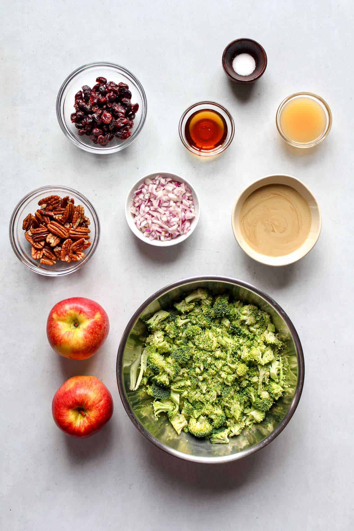 Overhead shot of all the individual ingredients for an Easy Vegan Broccoli Apple Salad, including broccoli, apples, pecans, cranberries, and tahini dressing components.