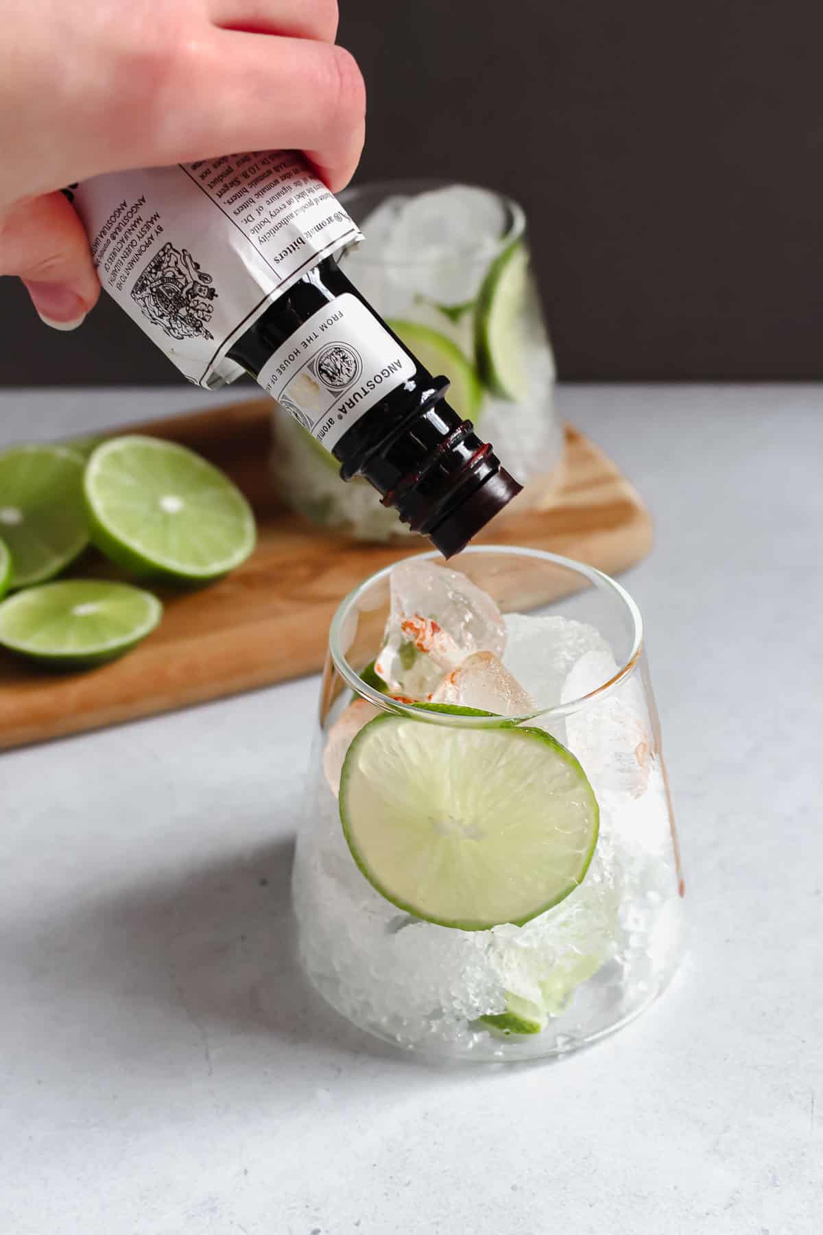 A hand shaking a bottle of bitters in to a glass that contains sliced limes and ice