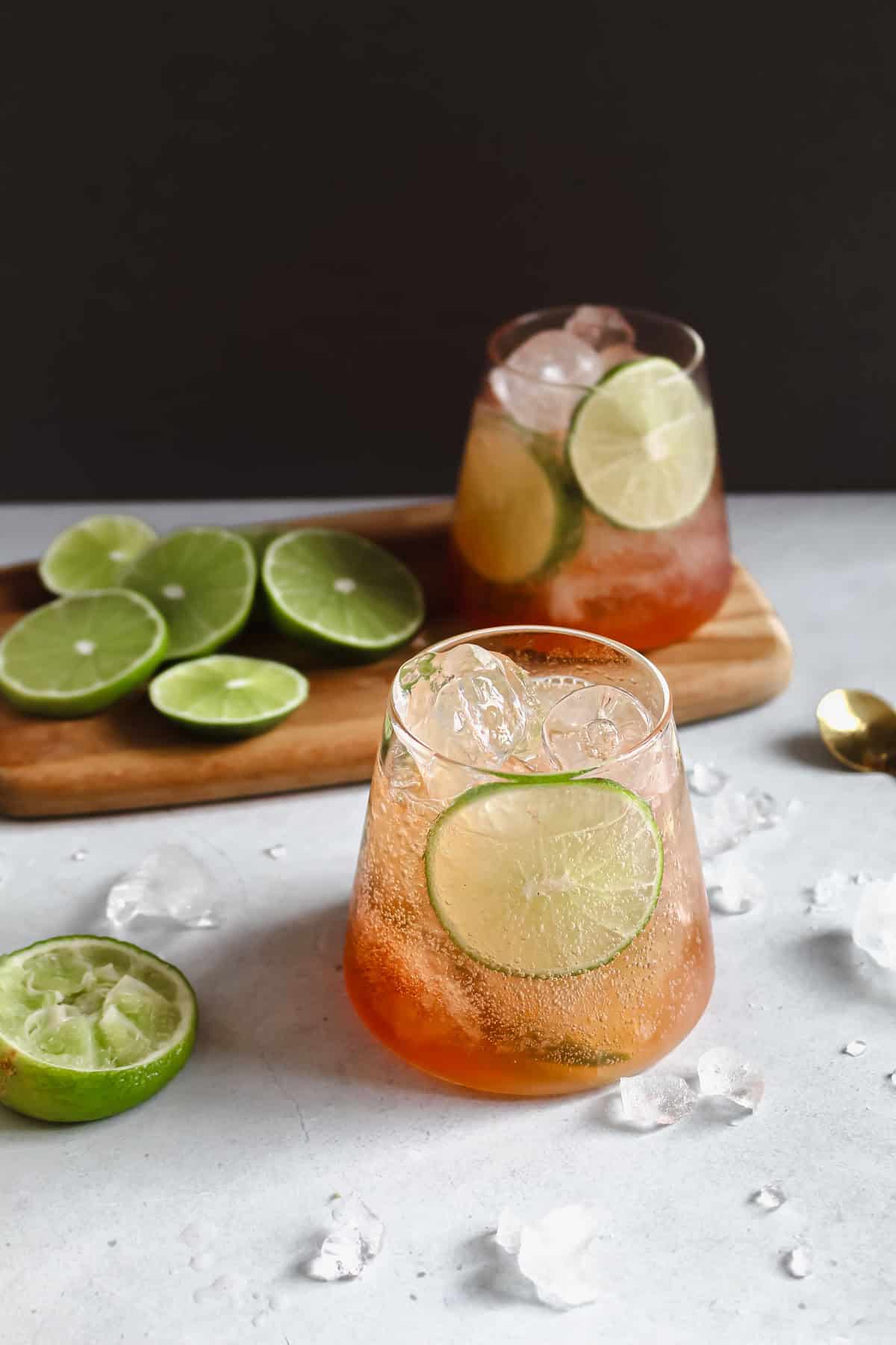 2 glasses filled with soda water, limes, and bitters accompanied by sliced limes on a wooden serving platter