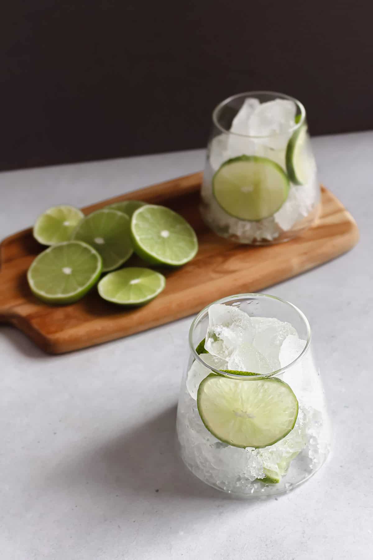 Ice and lime slices in a glass, accompanied by more sliced limes on a wooden serving board.