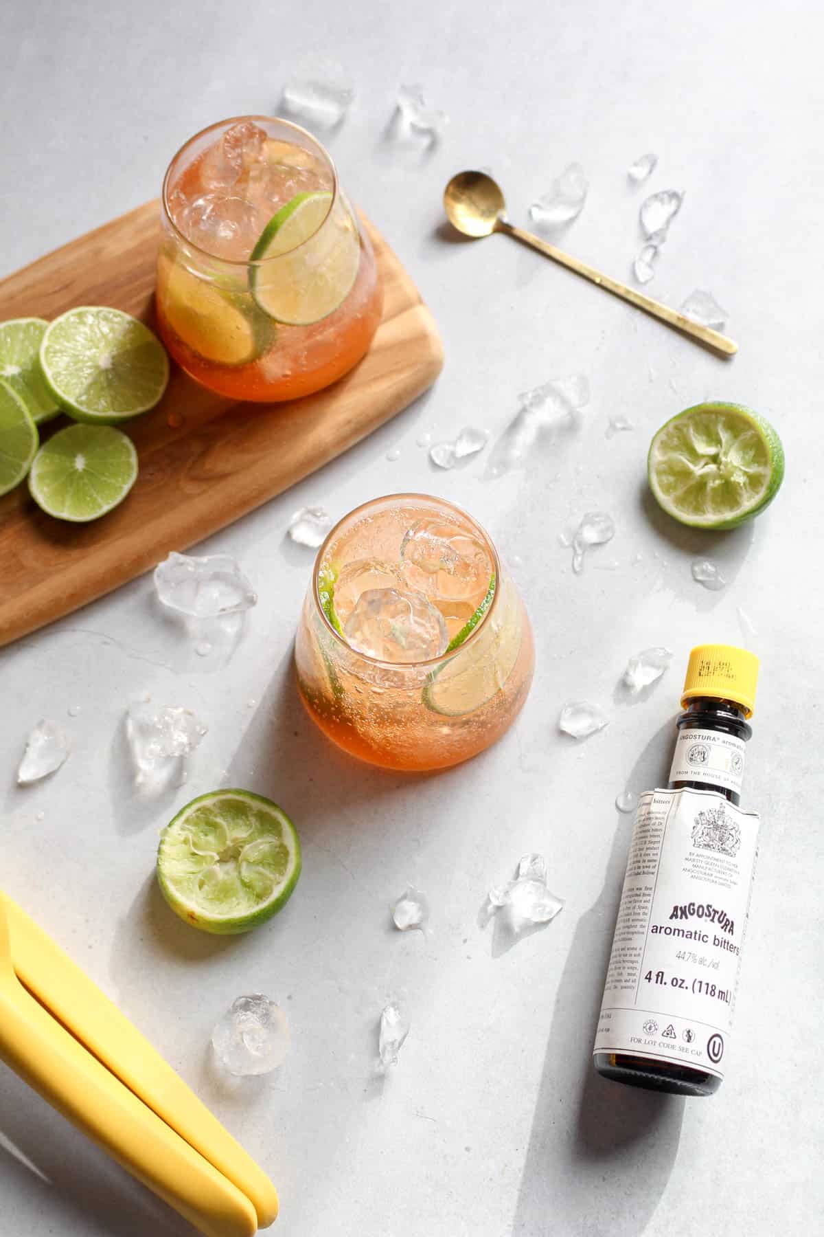 Two glasses of soda, lime, and bitters on a white background, accompanied by a bottle of bitters, fresh lime slices, and a golden stir spoon.