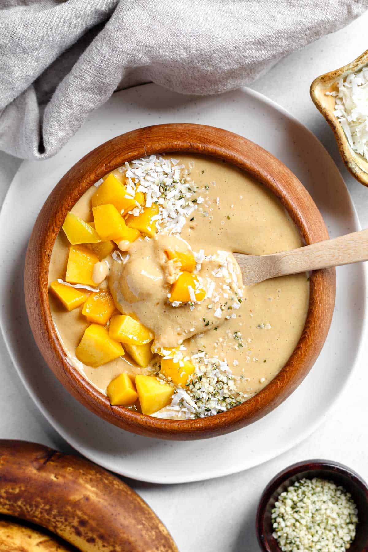 Spoon in creamy, high protein mango banana smoothie bowl that is topped with mango, shredded coconut, and hemp hearts.
