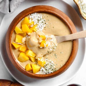Spoon in creamy, high protein mango banana smoothie bowl that is topped with mango, shredded coconut, and hemp hearts.