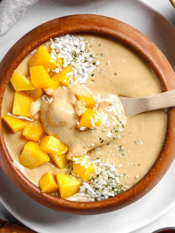Spoon in creamy, high protein mango banana smoothie bowl that is topped with mango, shredded coconut, and hemp hearts.