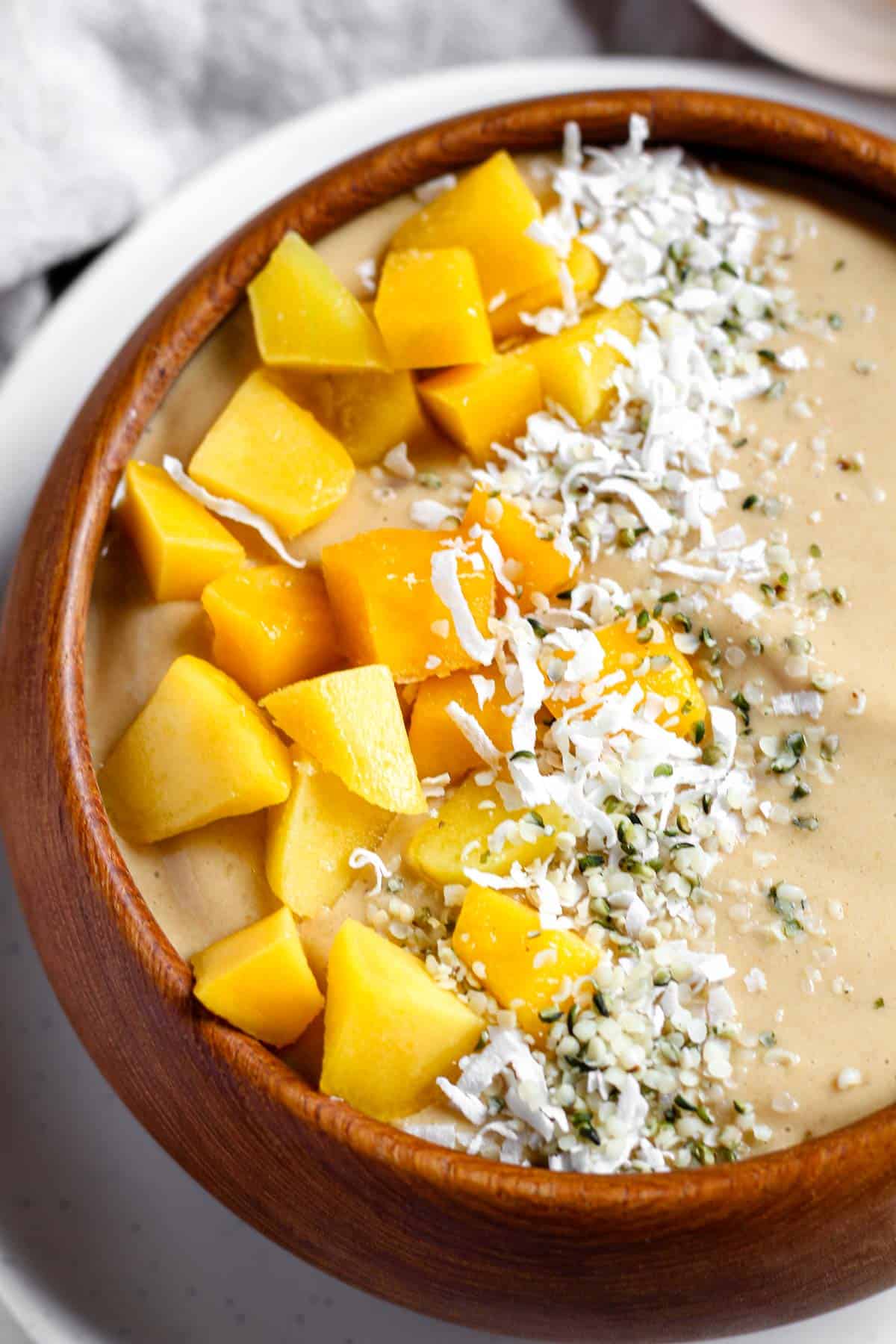 Close-up of a smoothie bowl topped with fresh mango chunks, shredded coconut, and hemp hearts, served in a wooden bowl.