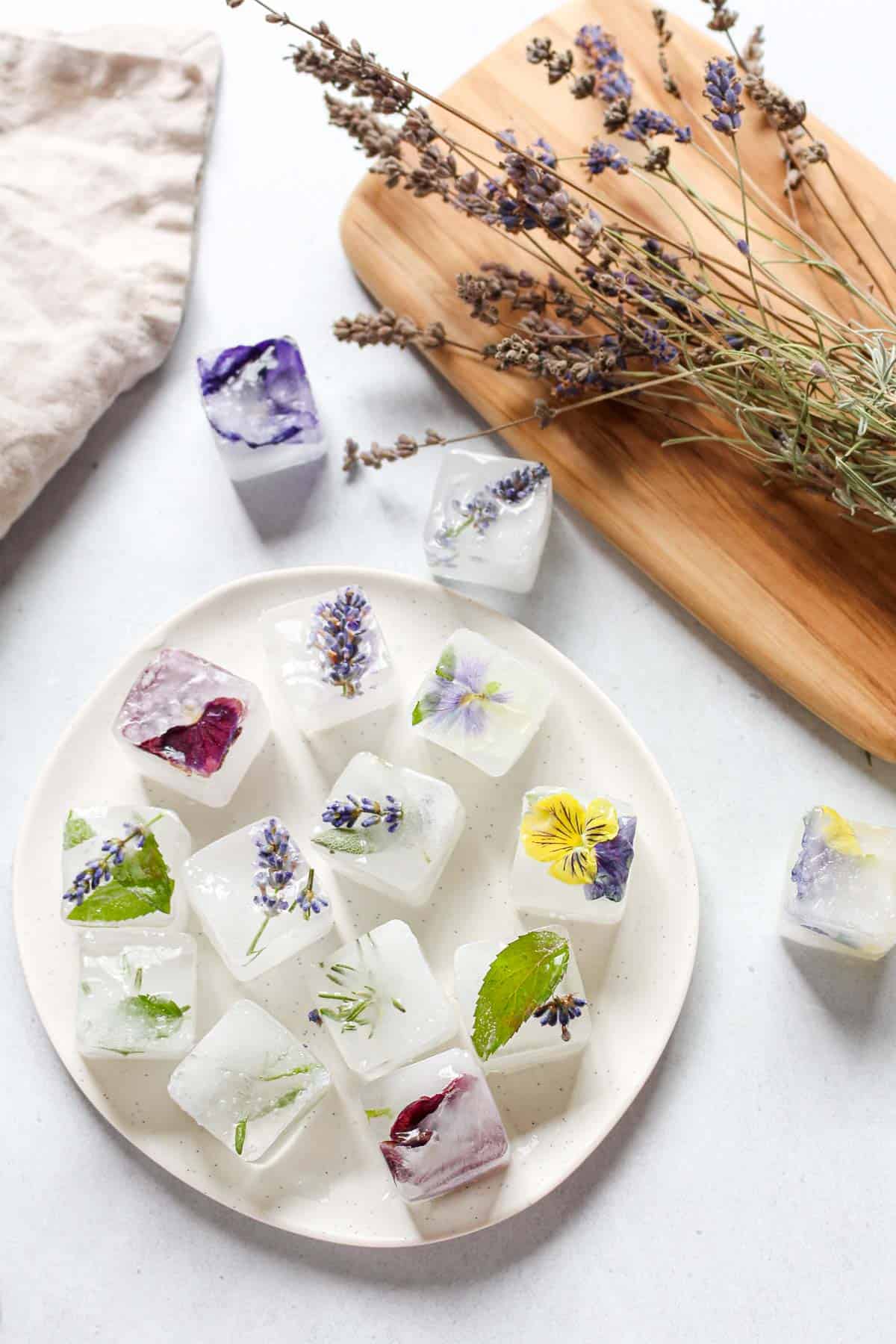 Ice cubes with flowers and herbs in them on a white plate next to dried lavender on wooden cutting board.