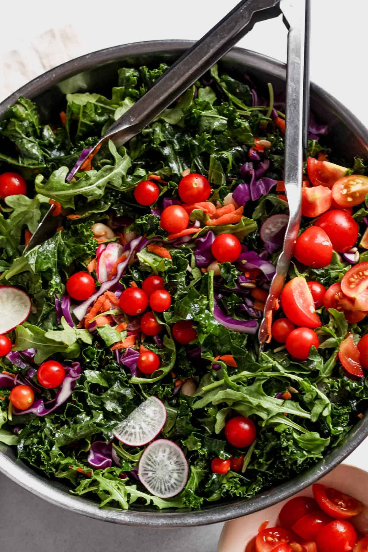 A stainless steel bowl filled with salad greens, purple cabbage, shredded carrots, and tomatoes with serving tongs.