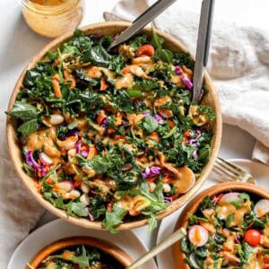 Kale and arugula salad in large bowl alongside two smaller wooden serving bowls filled with salad on a white background.