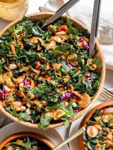 Kale and arugula salad in large bowl alongside two smaller wooden serving bowls filled with salad on a white background.