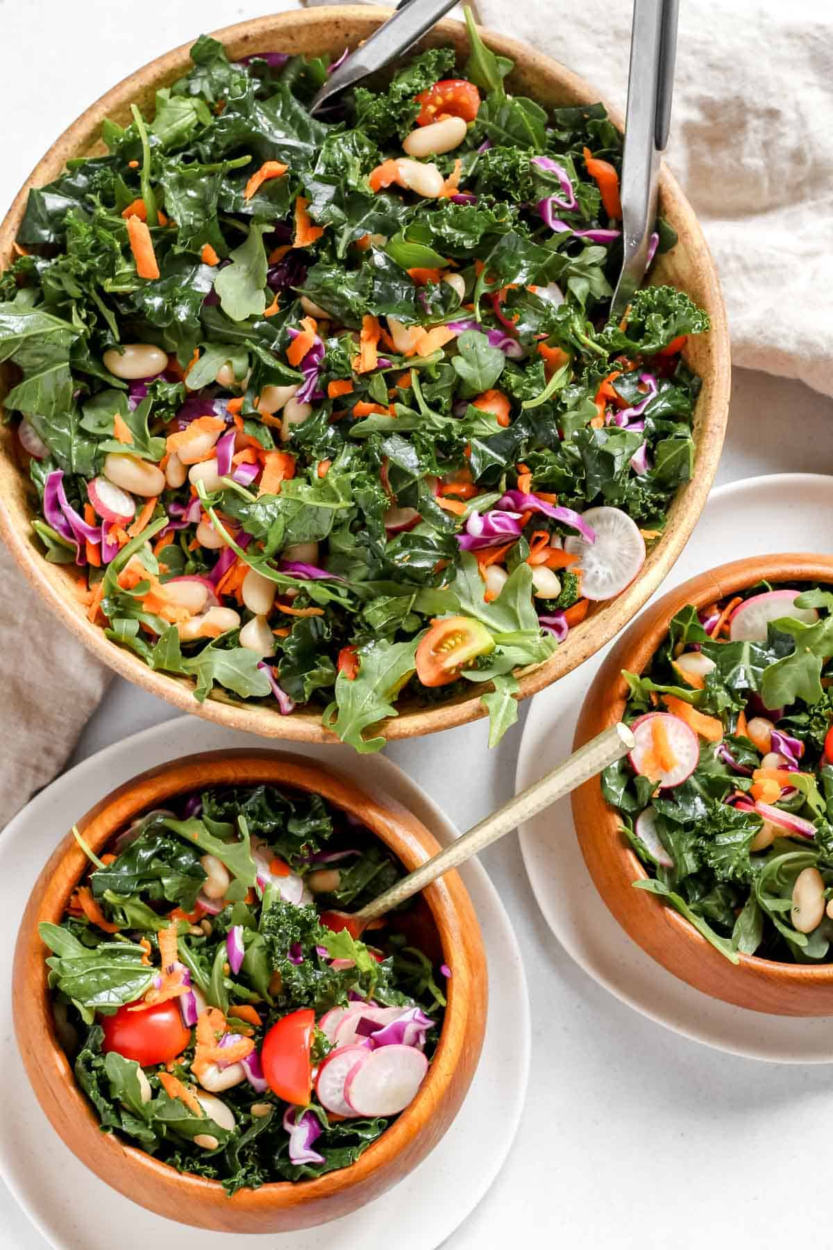 One large bowl full of kale and arugula salad alongside two smaller wooden serving bowls filled with salad on a white background.