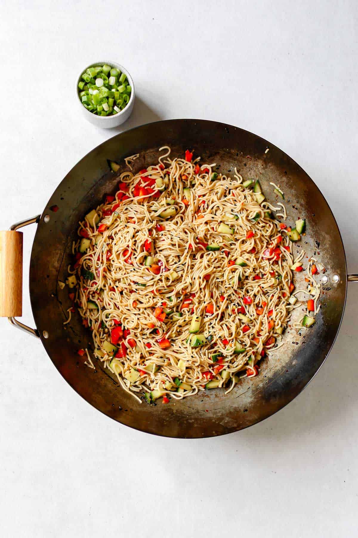 Steel wok with noodles, cucumber, and red bell pepper in it. A small bowl of green onions is next to the wok.