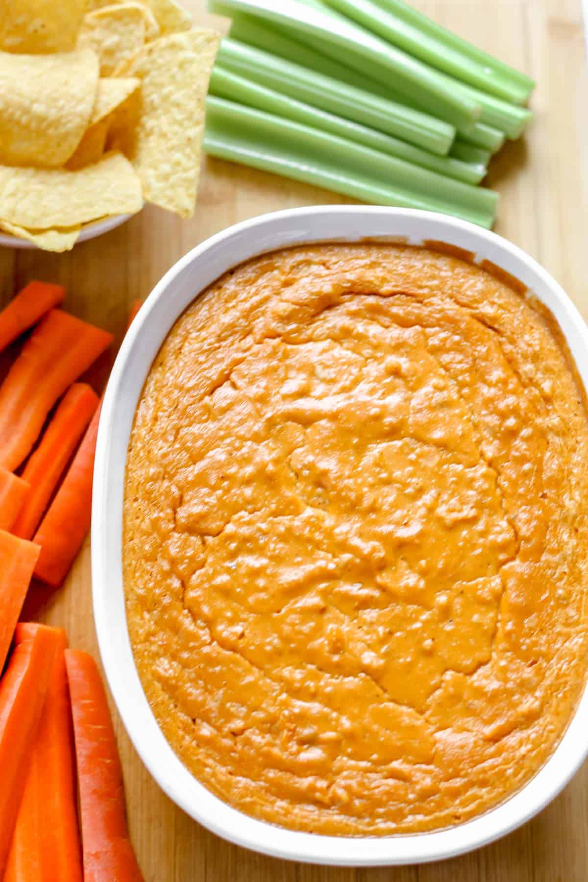 Baked vegan buffalo chicken dip in white baking dish on cutting board surrounded by celery, carrots, and tortilla chips